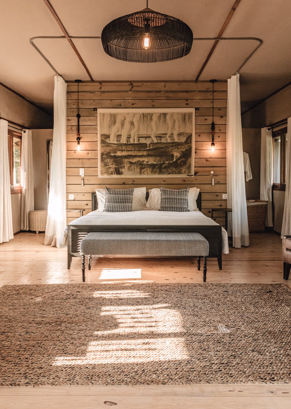 Cozy rustic bedroom featuring a canopy bed, a large nature tapestry, a black wicker chandelier, and a woven floor rug.