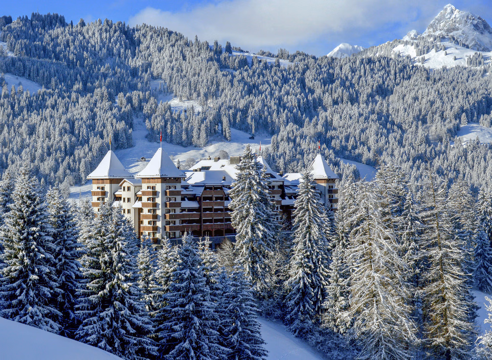 The Alpina Gstaad luxury hotel surrounded by snow-covered evergreen trees and mountains during winter.