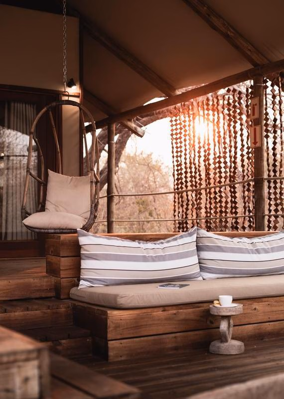 Shaded wooden deck at Toka Leya Camp with a hanging chair, daybed with striped pillows, and a small stool holding a cup.