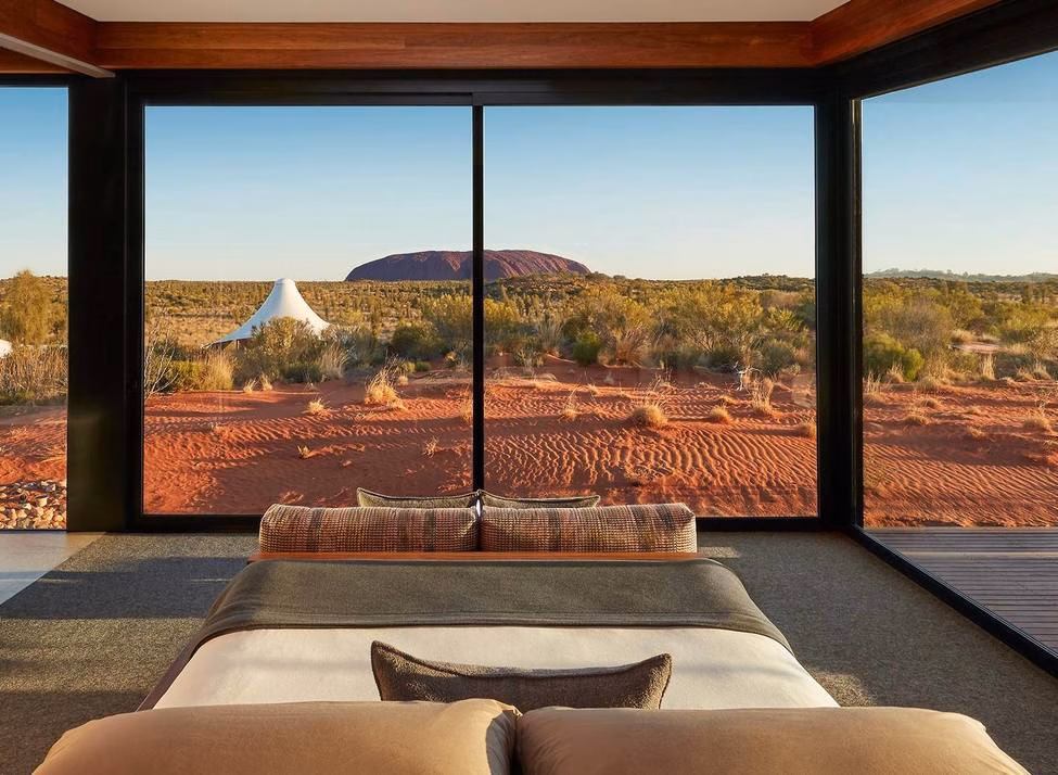 Bedroom with large floor-to-ceiling windows overlooking a red sand desert landscape and distant rock formation.