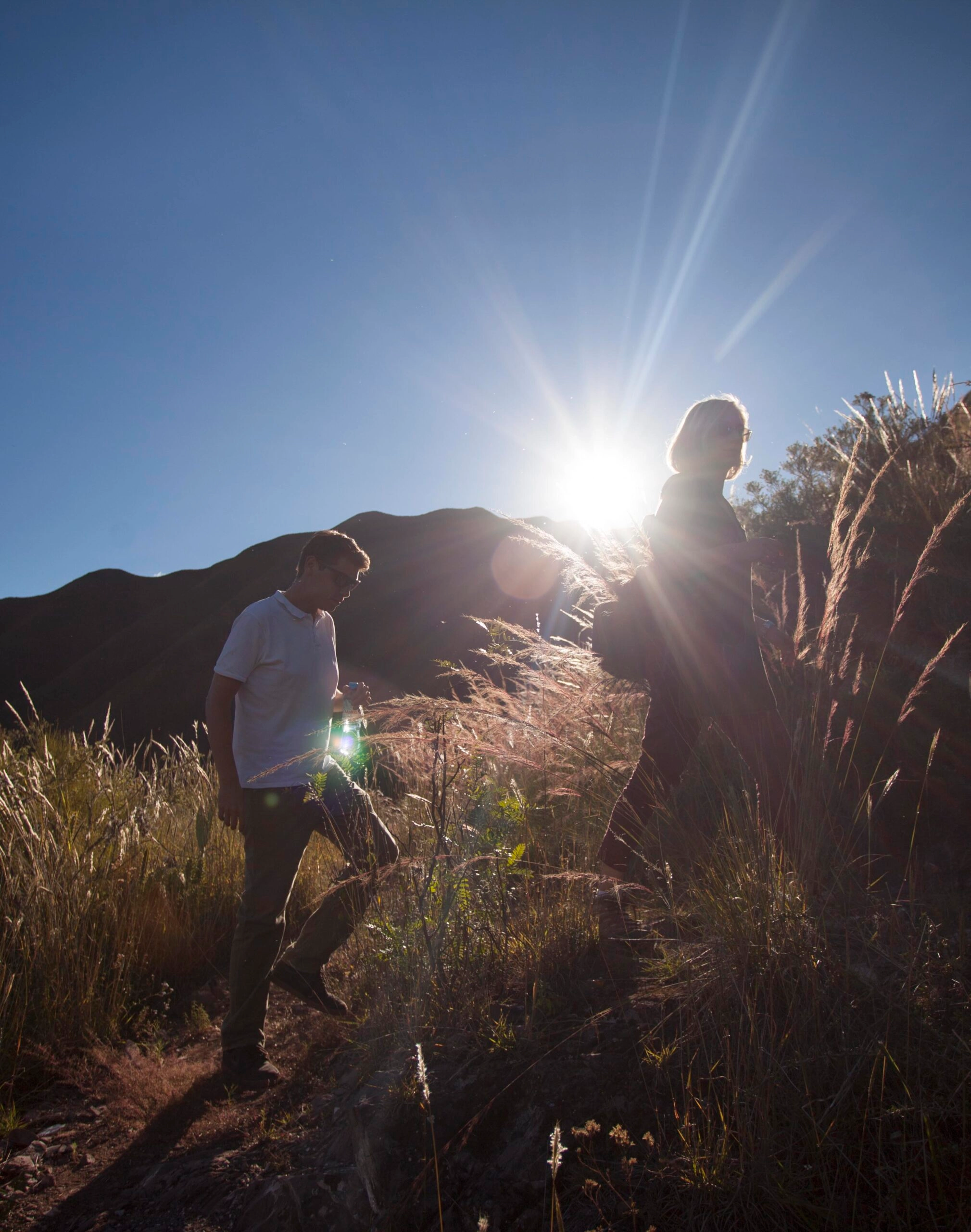 Two individuals hiking on a grassy trail with the sun low on the horizon.