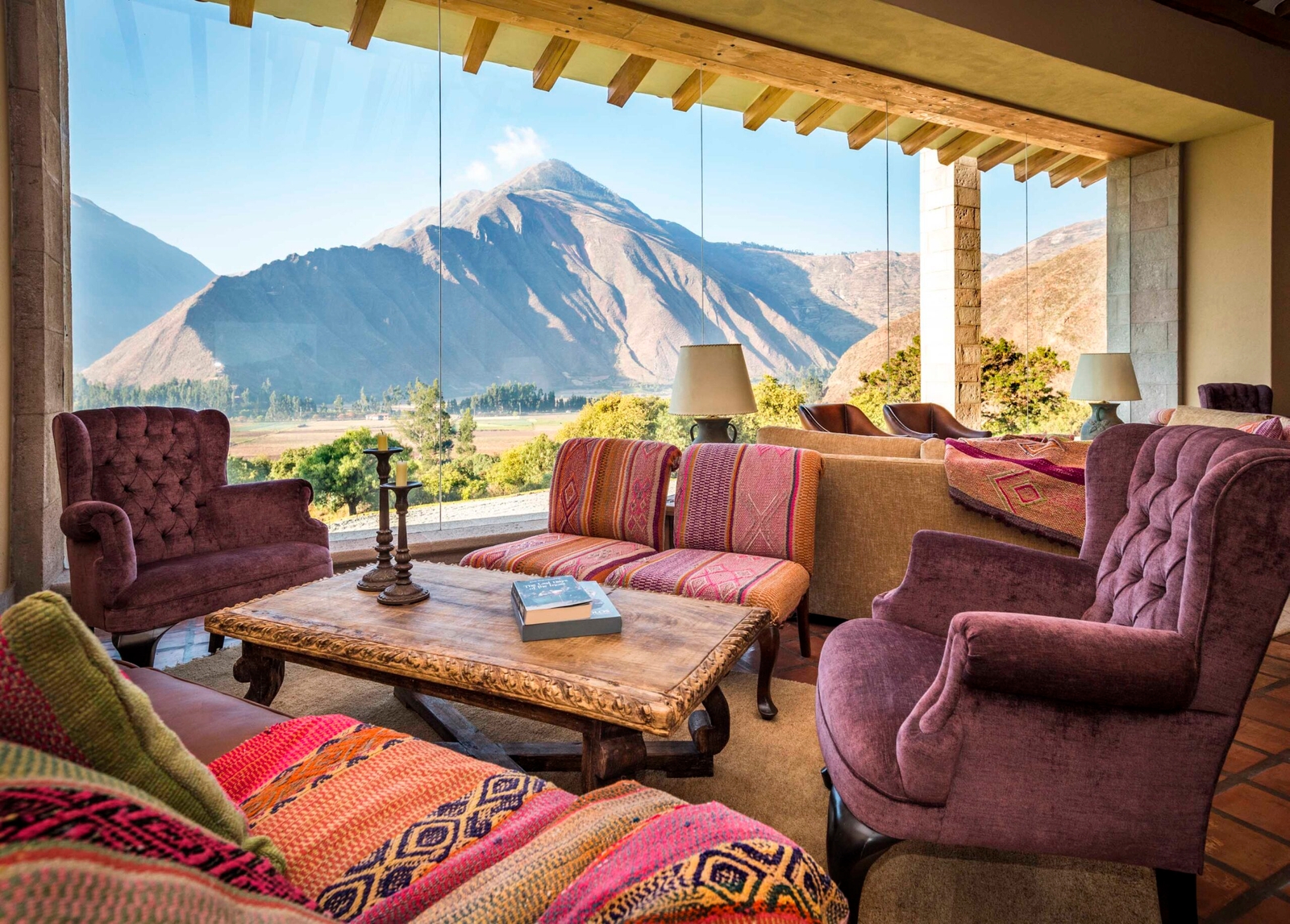 Cozy room with plush chairs, patterned cushions, rustic table, and a mountain view through a large window.