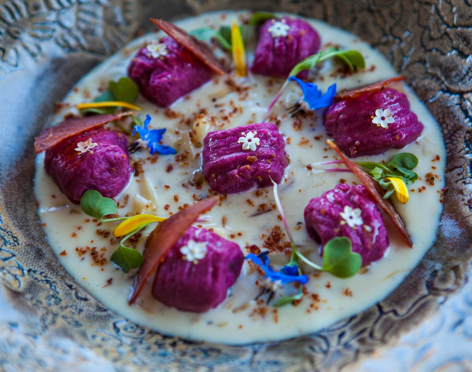 Gourmet dish with purple gnocchi, cream sauce, edible flowers, and herbs on ornate plate.