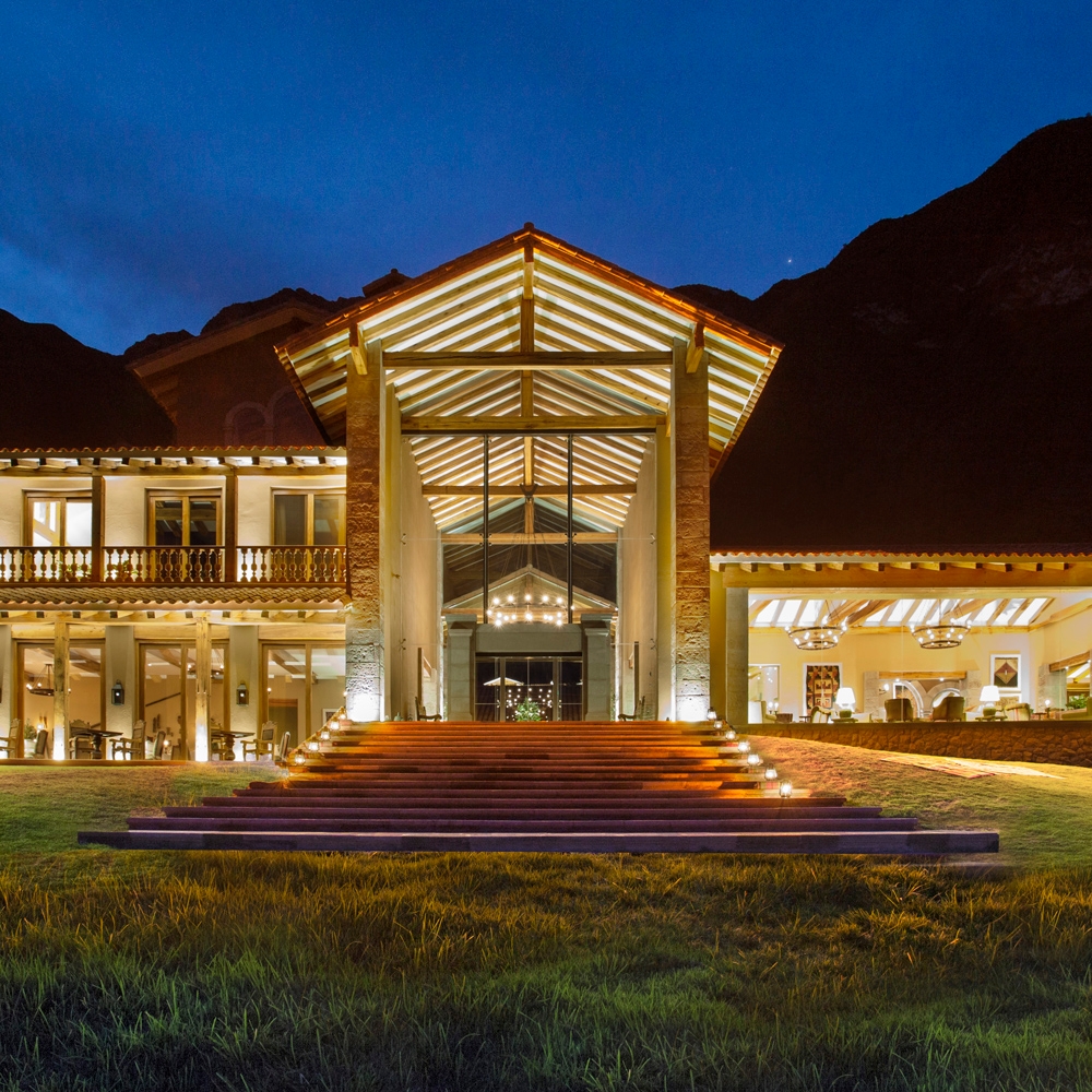 Illuminated facade of a luxury hotel at twilight with mountains in the background.