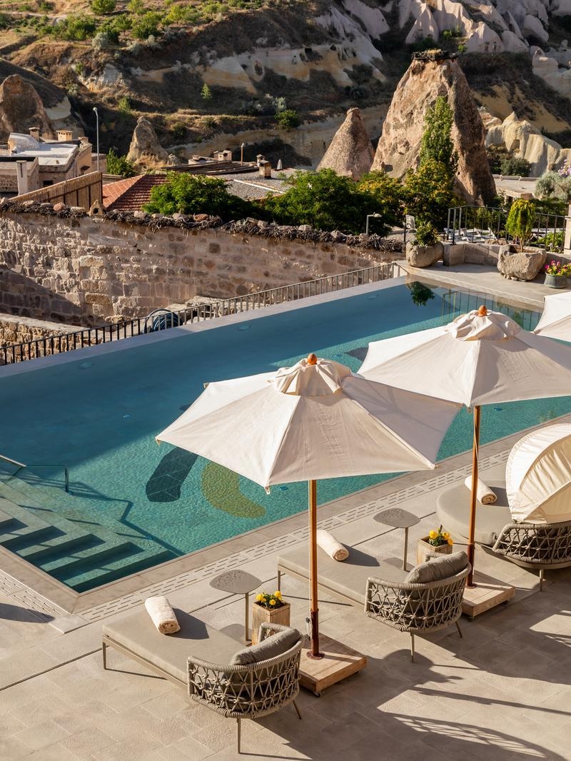 A blue infinity pool with sun loungers and umbrellas overlooking a rugged Turkish landscape.