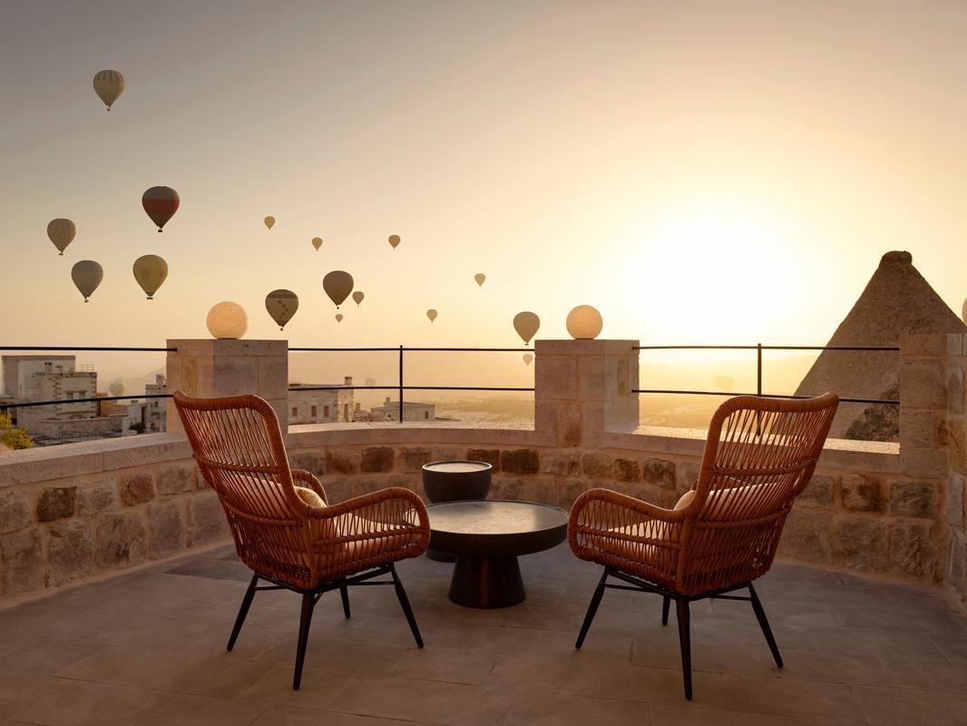 Wicker chairs on a stone terrace overlooking hot air balloons at sunrise in Cappadocia, Turkey.