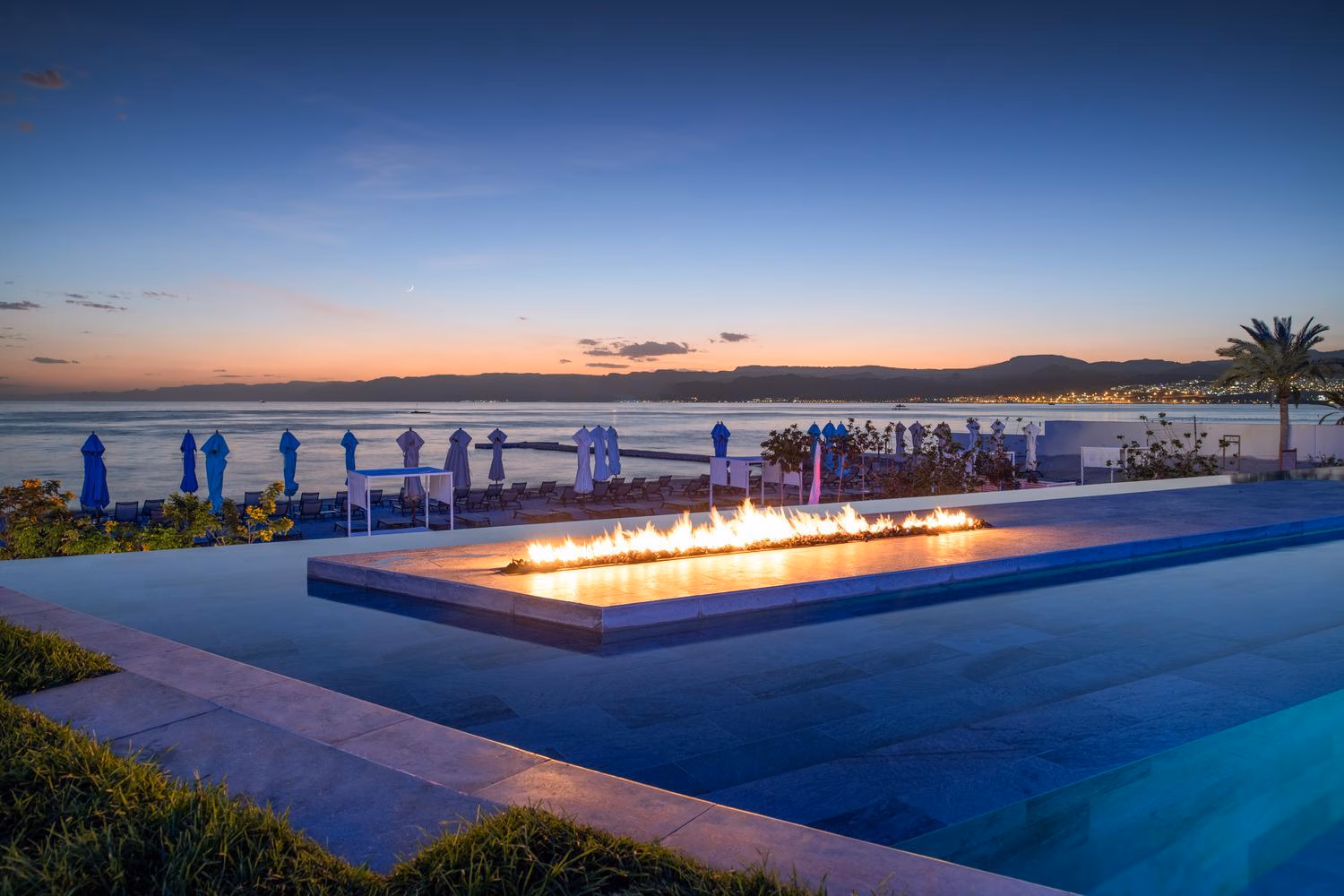 A long fire pit burning inside a modern infinity pool at twilight at Kempinski Hotel Aqaba, with sea views.