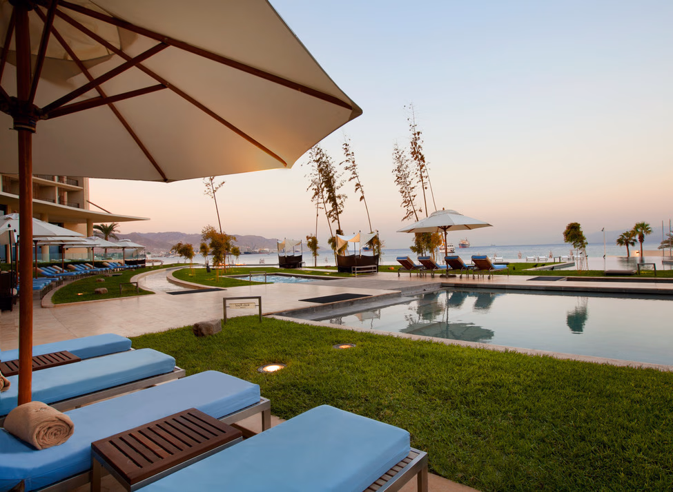 Blue lounge chairs and an umbrella by an outdoor pool at Kempinski Hotel Aqaba, overlooking the sea.