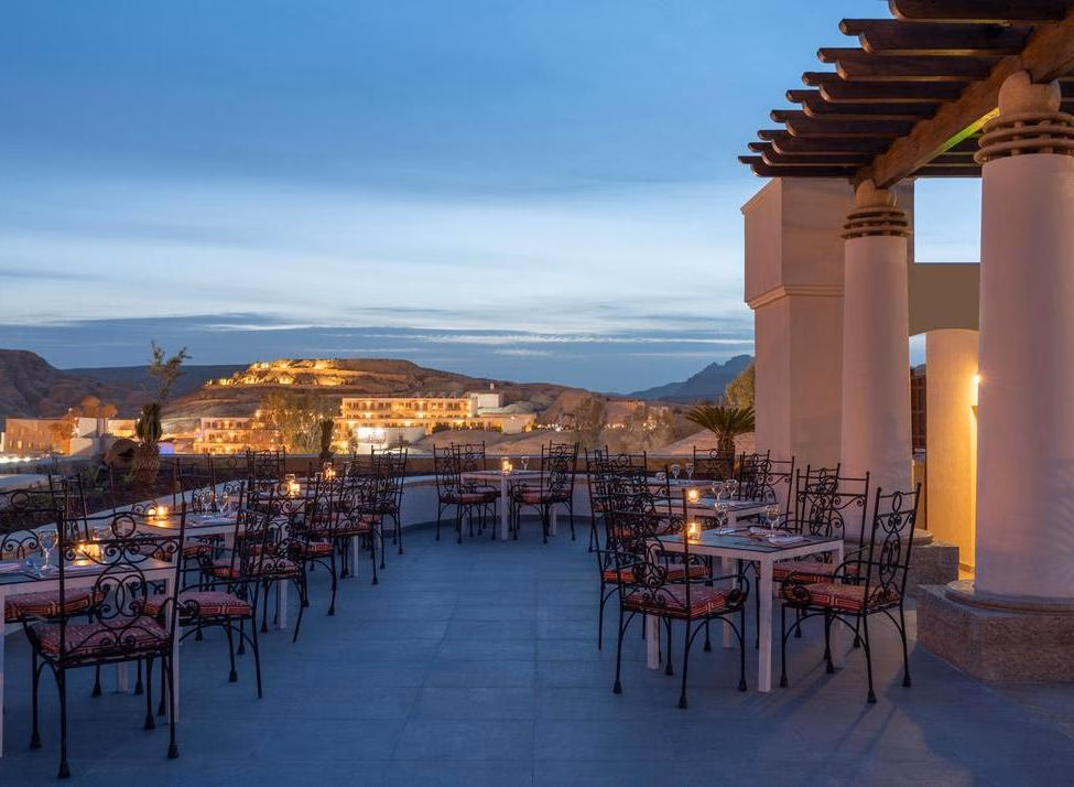 An outdoor restaurant terrace with set tables and candles overlooking a lit-up hillside town at dusk.