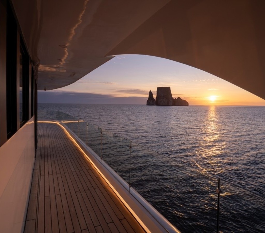 Teak wood yacht deck with glass railing framing a sunset over the ocean with rock formations on the horizon.
