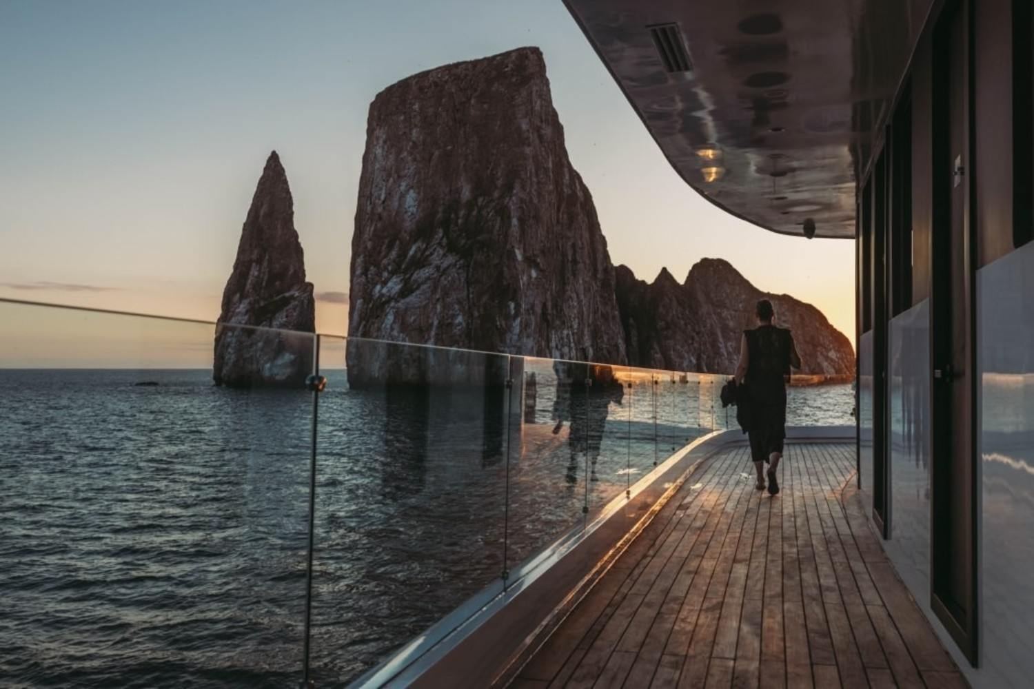 Person walking on a catamaran’s glass-railed wooden deck facing large rock pinnacles in the ocean.