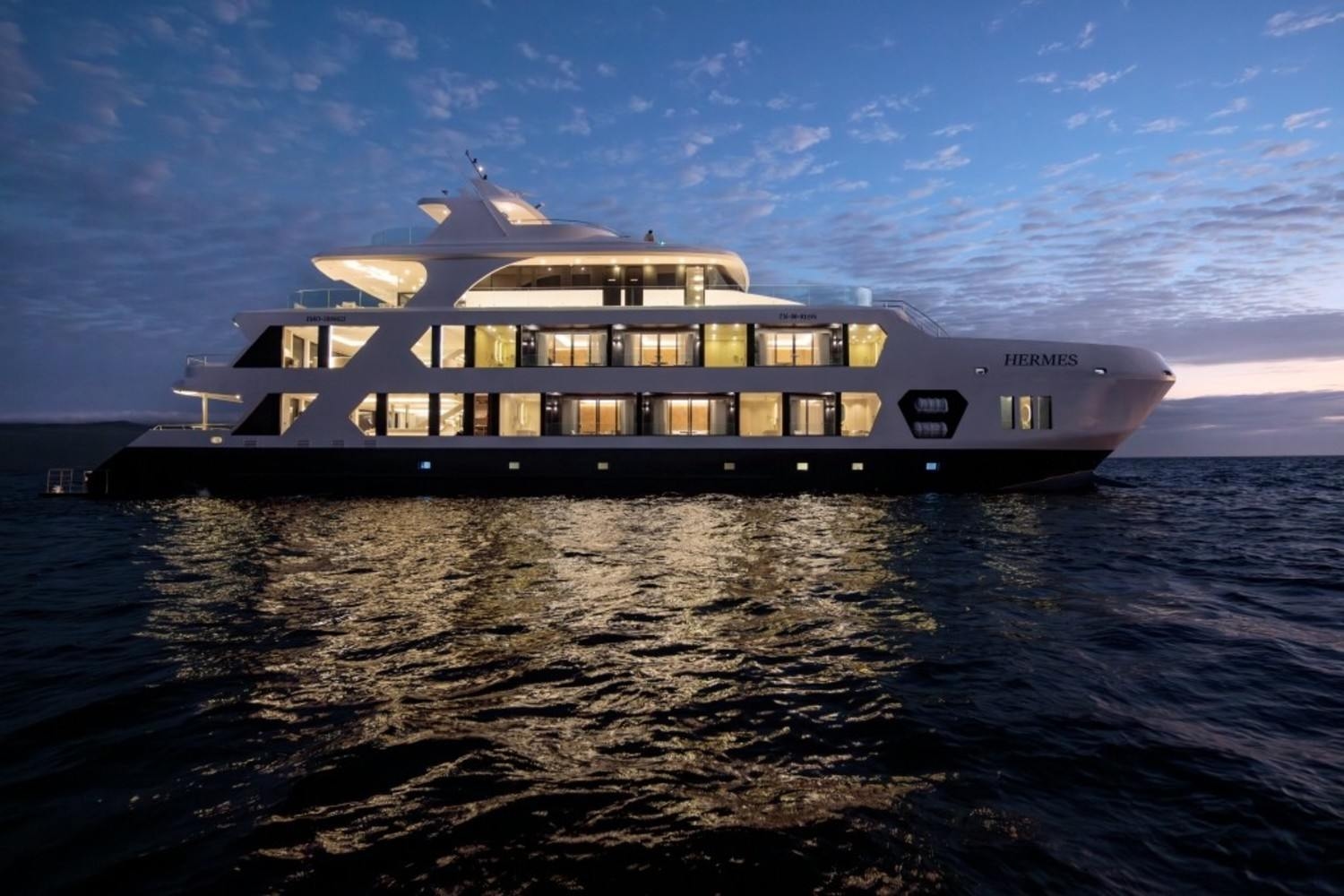 Profile view of the illuminated Hermes mega catamaran at sea during dusk.