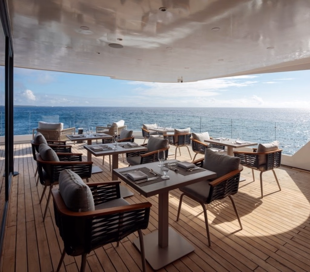 Covered wooden deck of a catamaran set with dining tables and chairs facing the ocean.