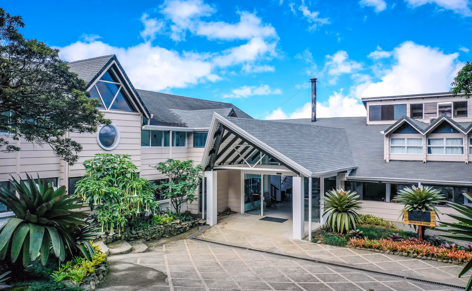 Exterior of Monteverde Lodge showing the white gabled entrance, stone pathways, and tropical plants.