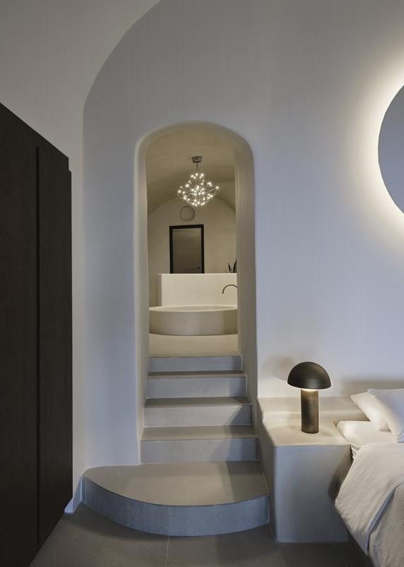 Archway with steps leading to a round soaking tub under a chandelier in a white bedroom at Andronis luxury suites.