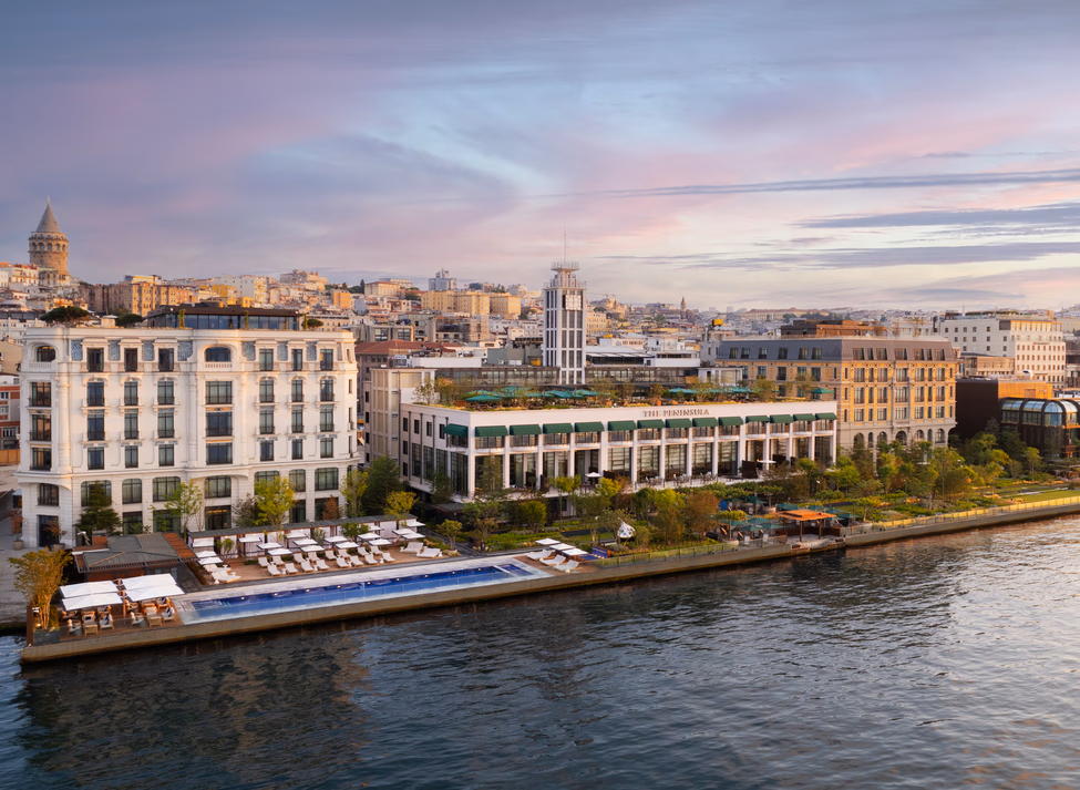 A wide aerial view of a luxury waterfront hotel with an outdoor pool, gardens, and a city skyline at sunset.