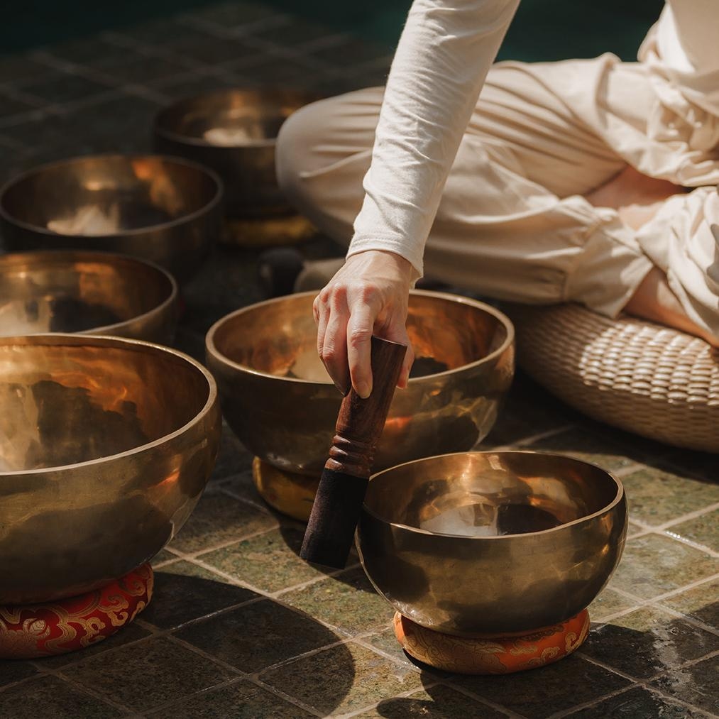 Person in white clothing playing a Tibetan singing bowl among several bowls.
