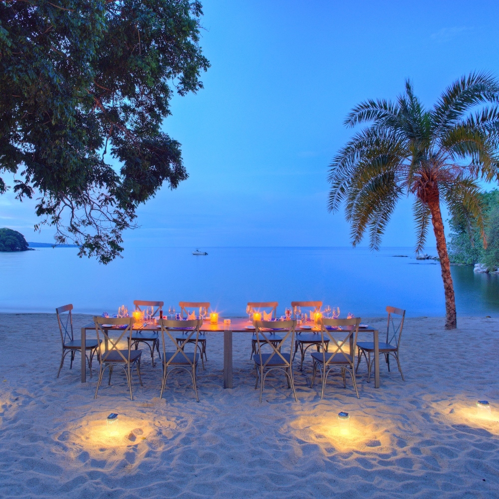 Beachside evening dining setup with lit candles on tables, palm trees, and calm sea background.