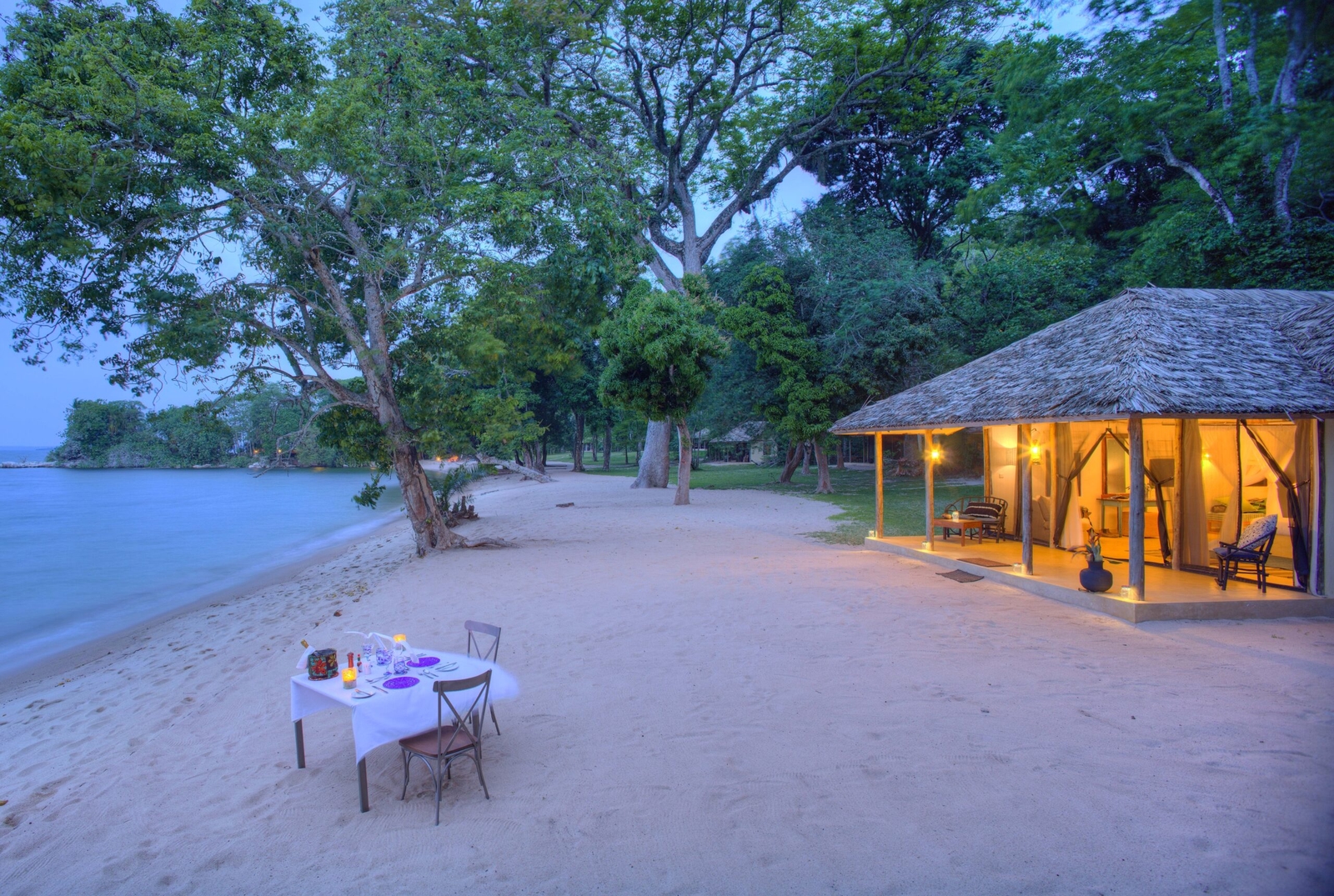Beachside dinner table setup near thatched-roof cabin at twilight.