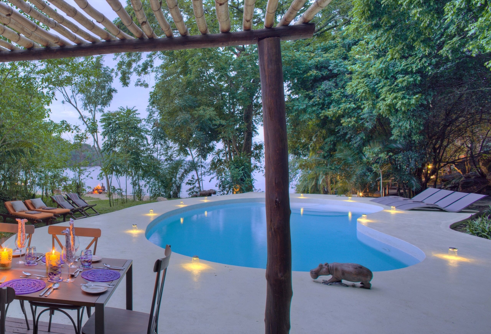 Outdoor dining setup by an infinity pool with lit candles and a forested backdrop at dusk.
