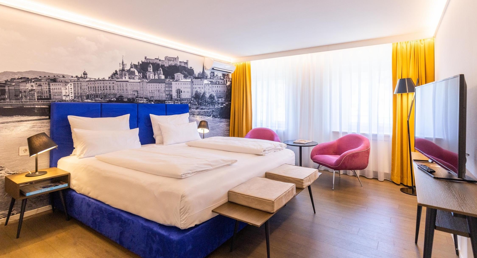 Modern hotel room with blue bed, wall-sized cityscape photo, pink chairs, yellow curtains, and flat-screen TV.