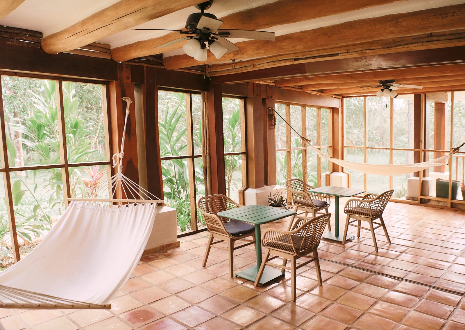 Cozy sunroom with hammock, dining set, ceiling fans, and lush greenery outside windows.