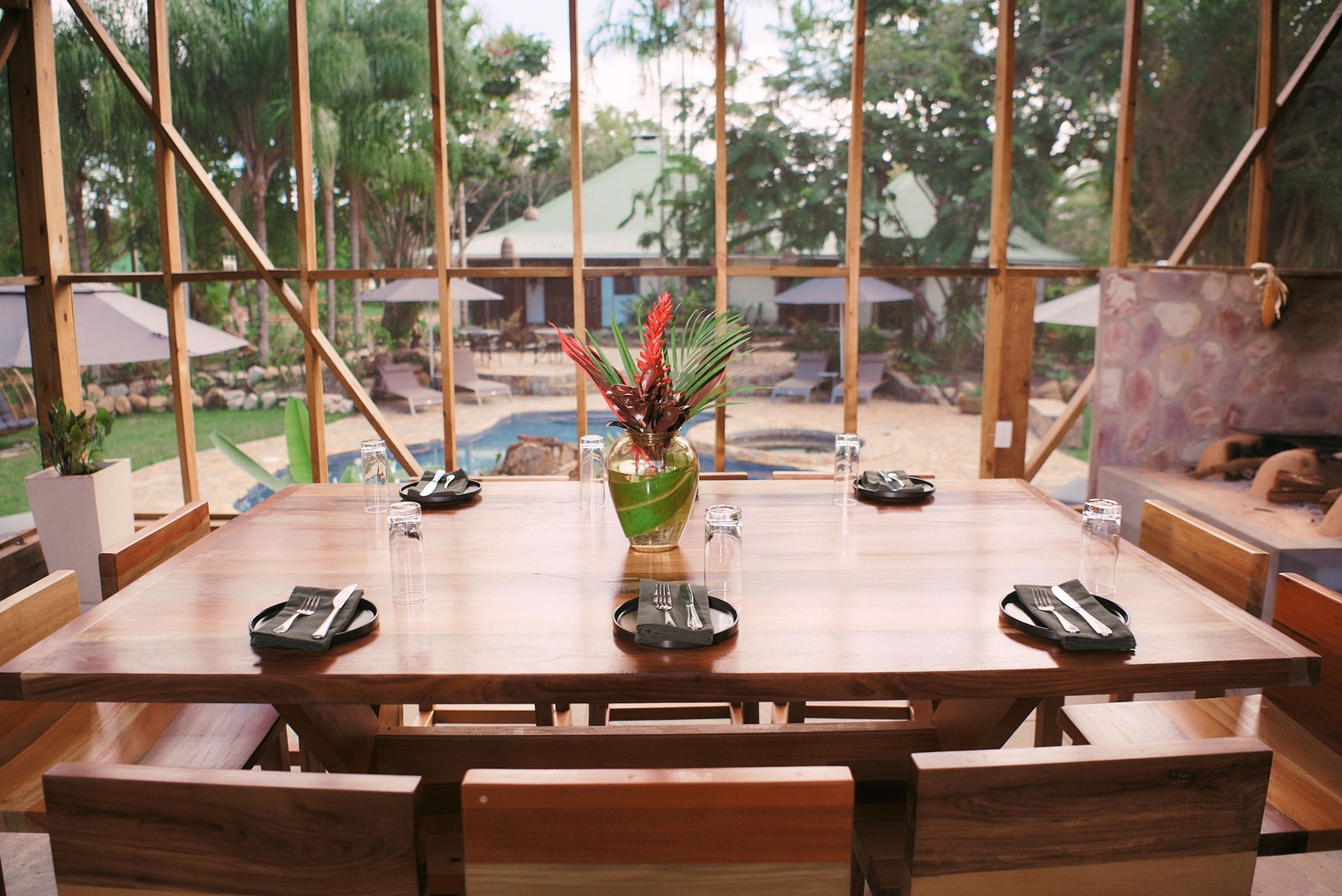 Wooden dining table with settings, overlooking a pool area through large windows.