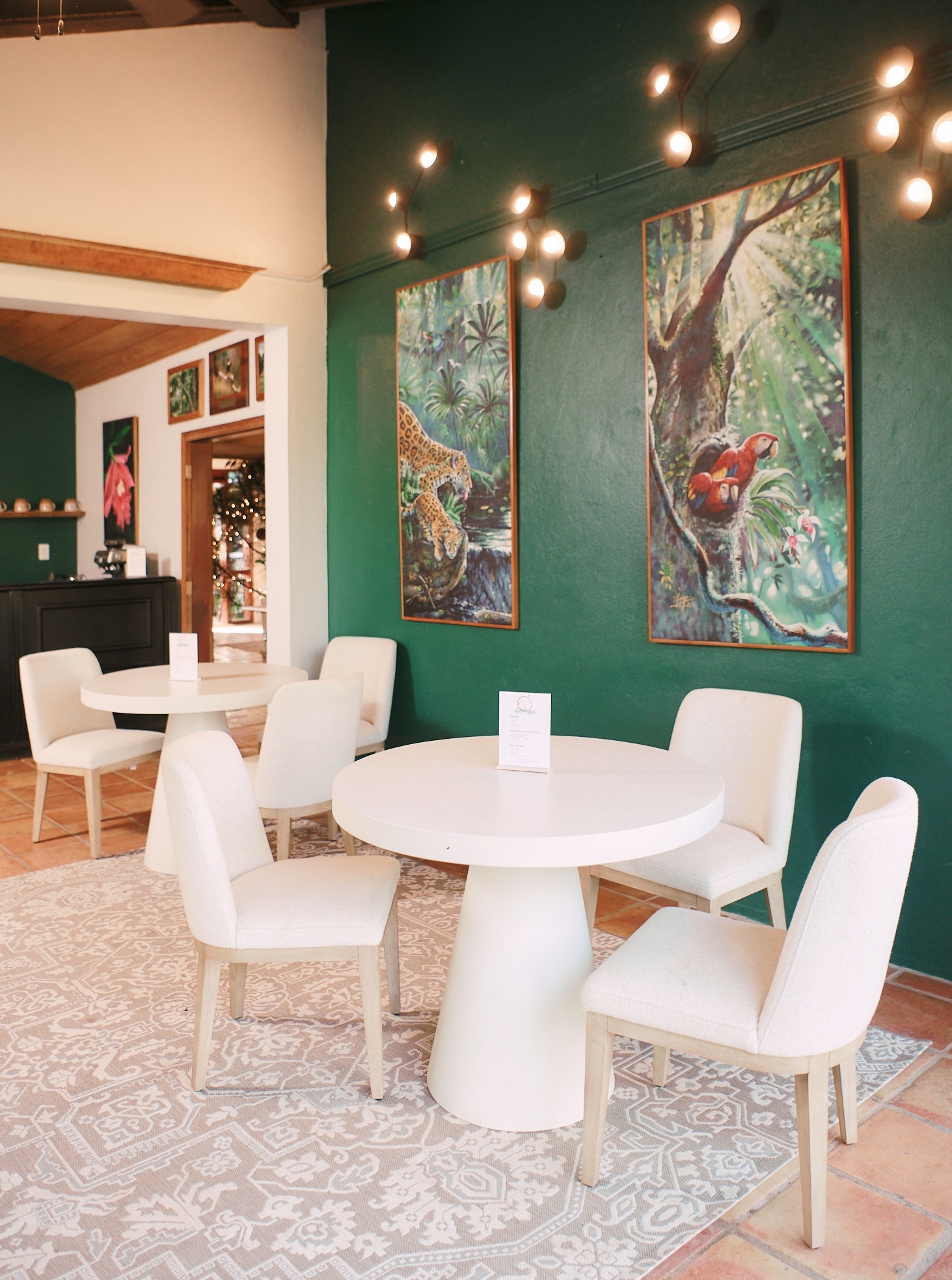 Interior with a round table, chairs, and wildlife paintings on a green wall, illuminated by string lights.