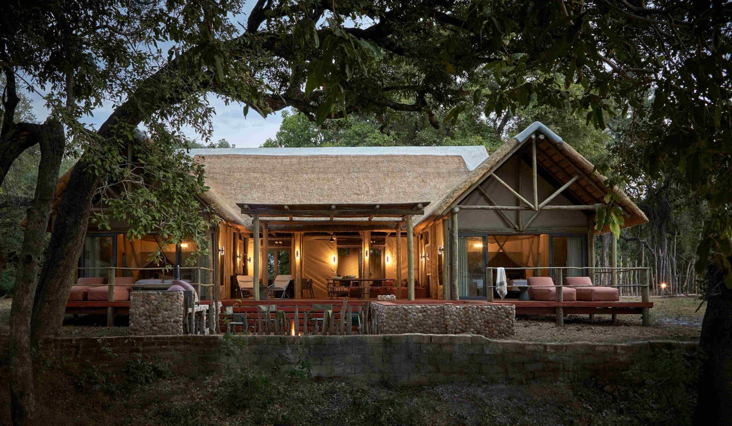 Thatched-roof lodge with lit interior surrounded by trees at twilight.