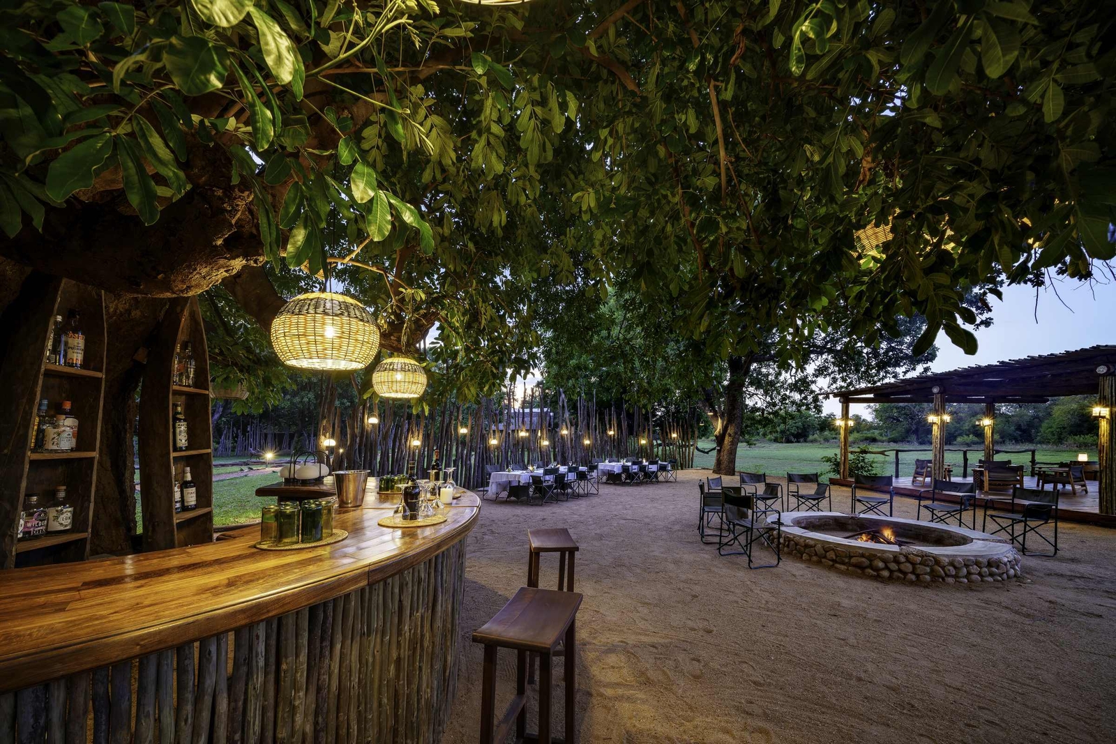 Outdoor dining space with bar, hanging lights, and a fire pit at dusk surrounded by trees.
