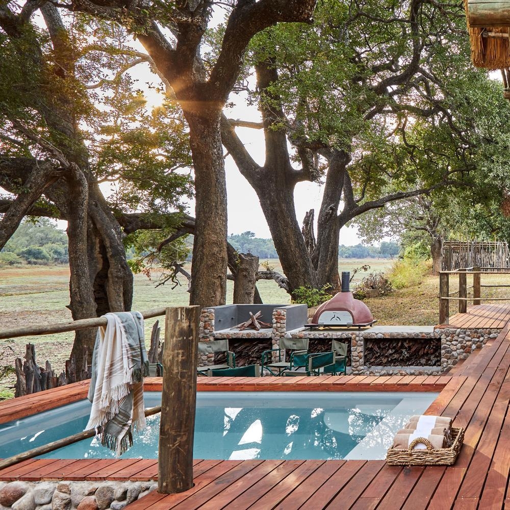 Rustic outdoor pool area with a pizza oven and seating surrounded by large trees.