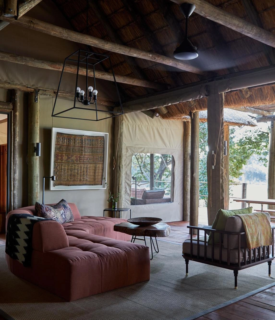 Rustic living room with thatched roof, large sofa, wooden beams, and rolled-up canvas walls.