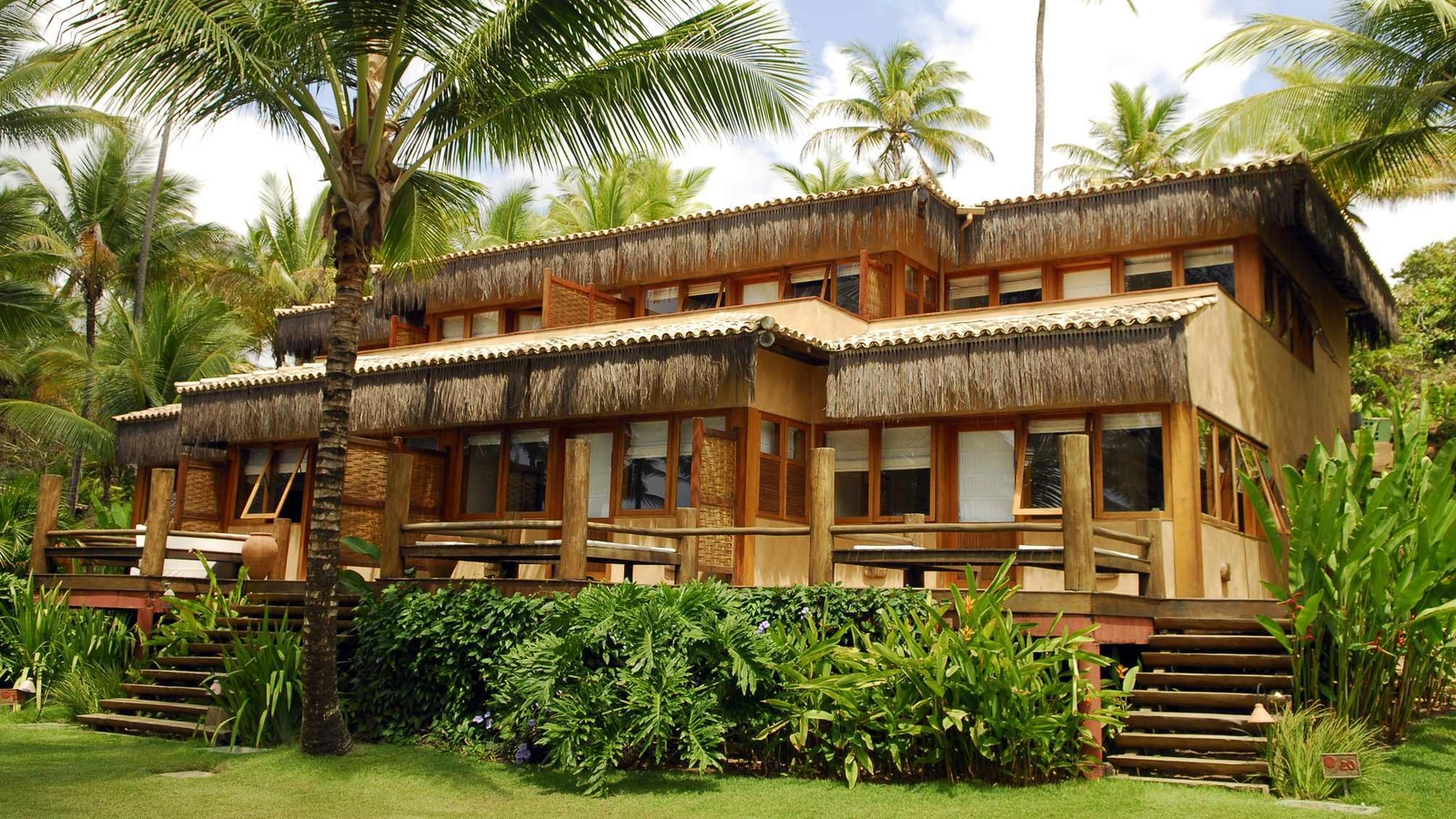 Tropical wooden house with thatched roof, surrounded by palm trees and lush greenery, featuring balconies and wooden stairs.