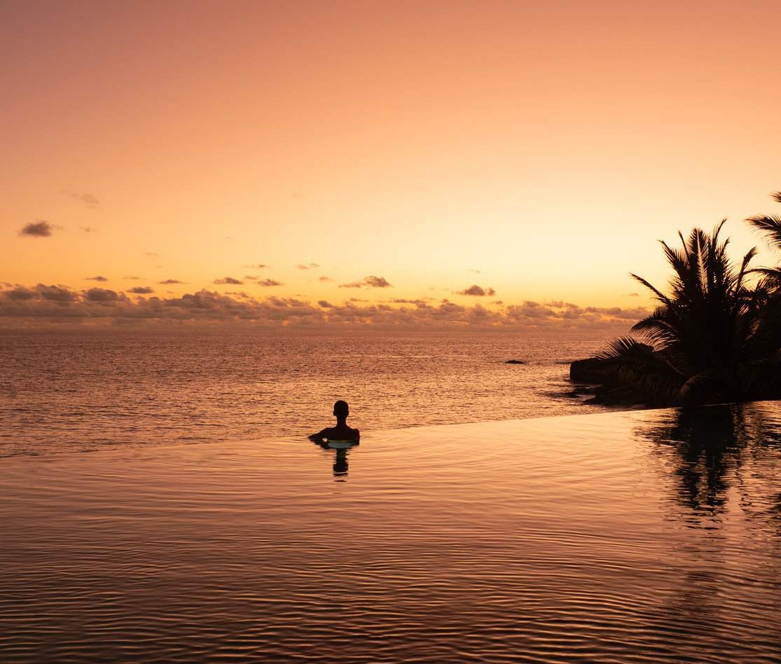 A woman looking to the horizon from an infinity pool at sunset, at Cheval Blanc Seychelles.