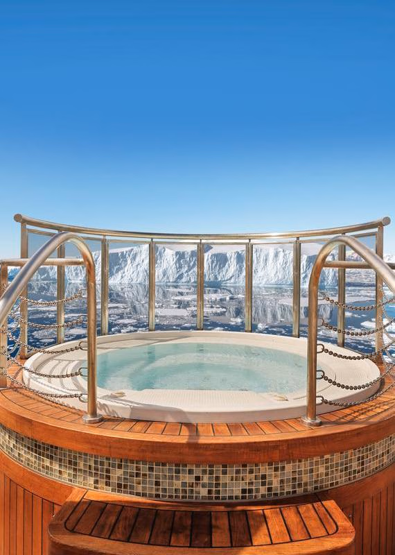Round hot tub on a wooden ship deck enclosed by glass railings, looking out at a massive iceberg in calm, icy waters.