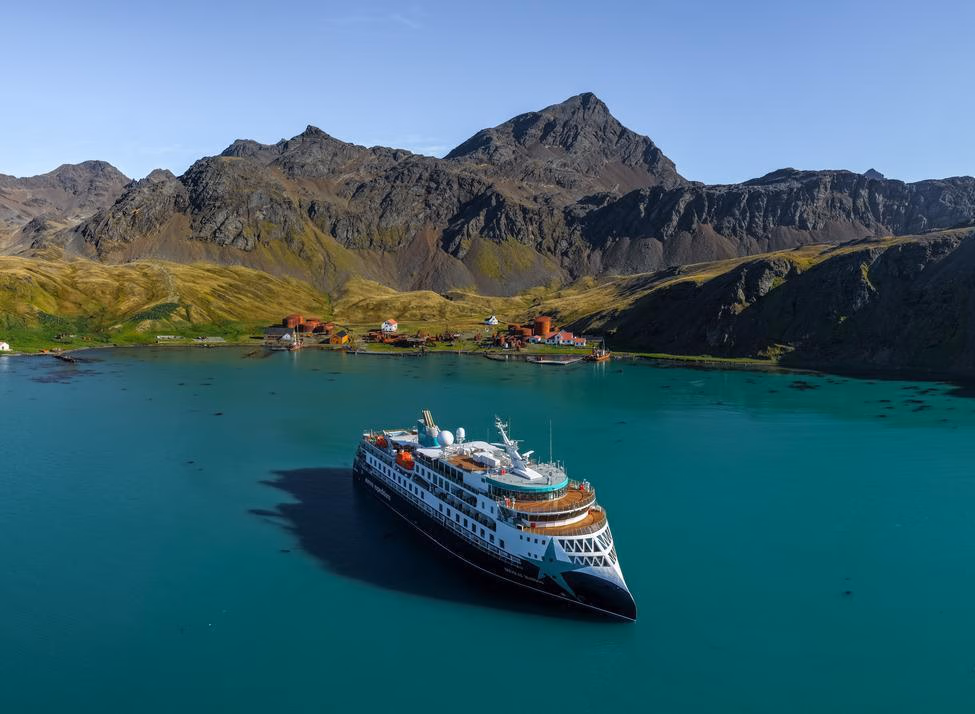 Large expedition cruise ship anchored in turquoise water near a shoreline with rusted buildings and rugged green mountains.
