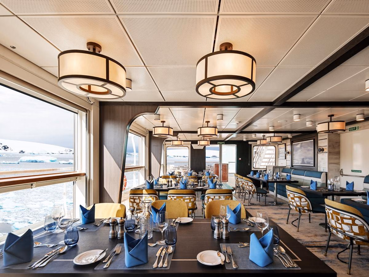Bright dining room with set tables, blue napkins, and large windows looking out at an icy seascape.