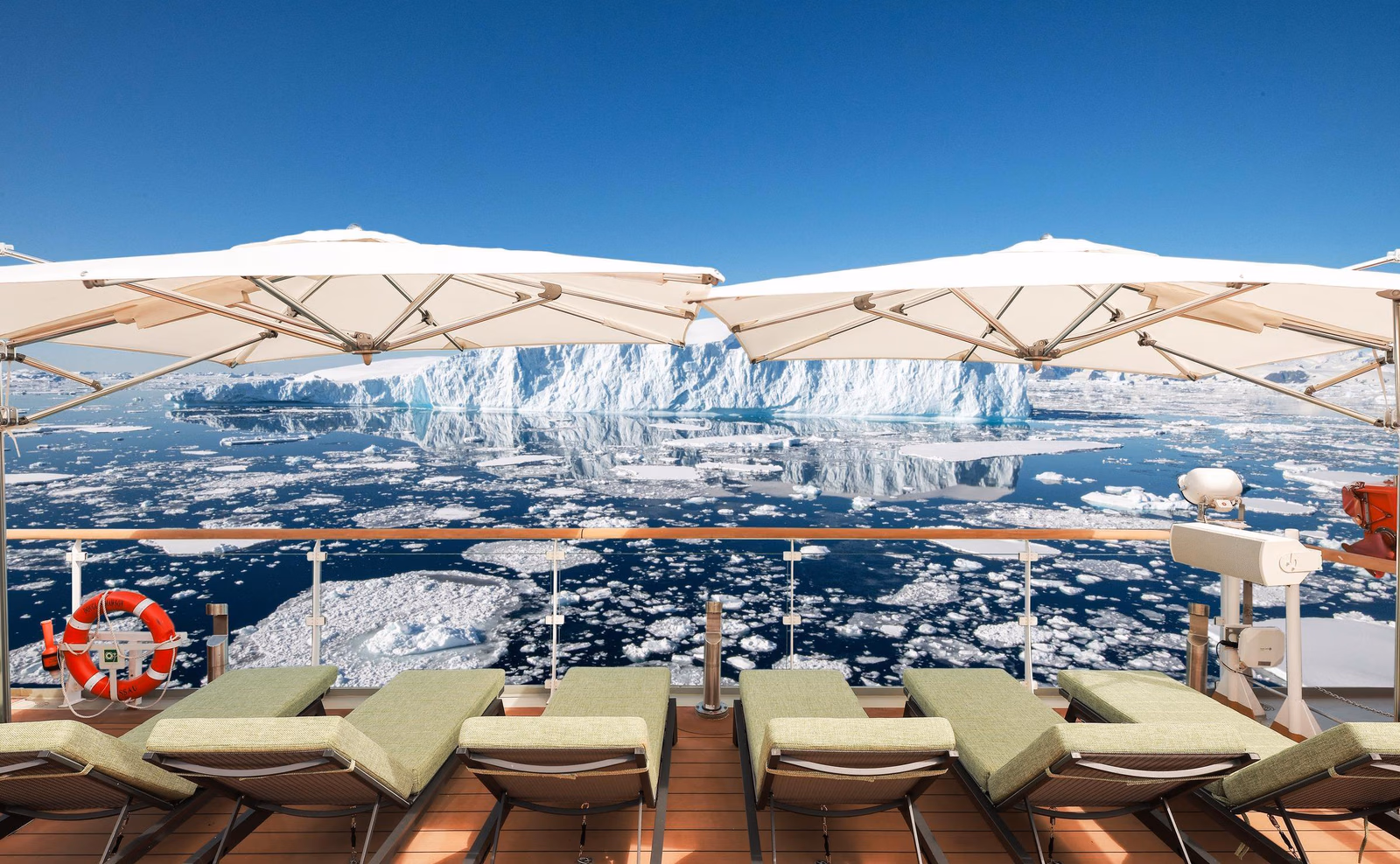 Ship deck with green lounge chairs, white umbrellas, and a life ring, facing a large iceberg surrounded by sea ice.