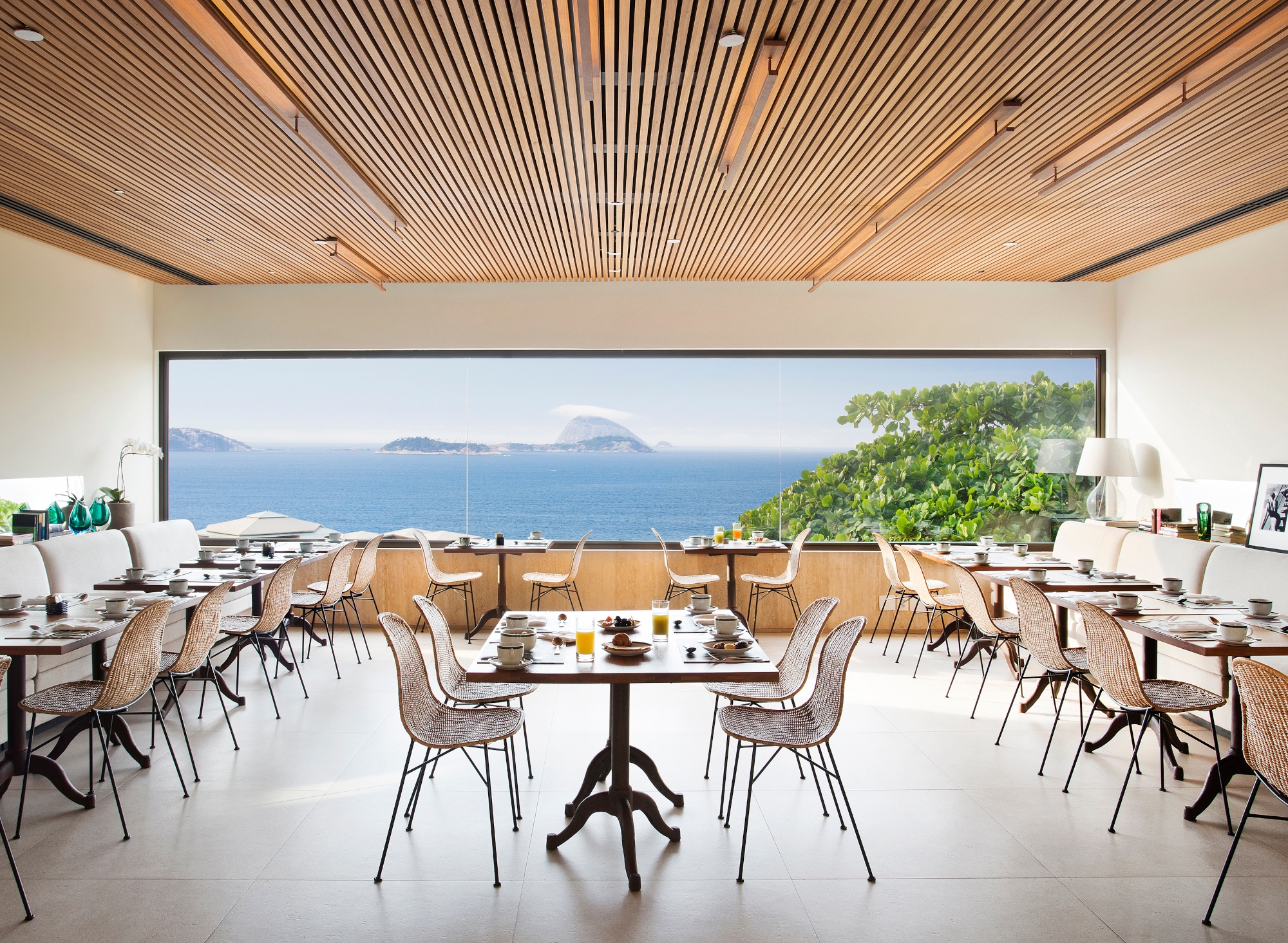 A dining room with rattan chairs and tables under a wooden ceiling and with a white floor. In the background is a long picture window looking out over some greenery and in the distance, the blue sea, some islands and the blue sky.