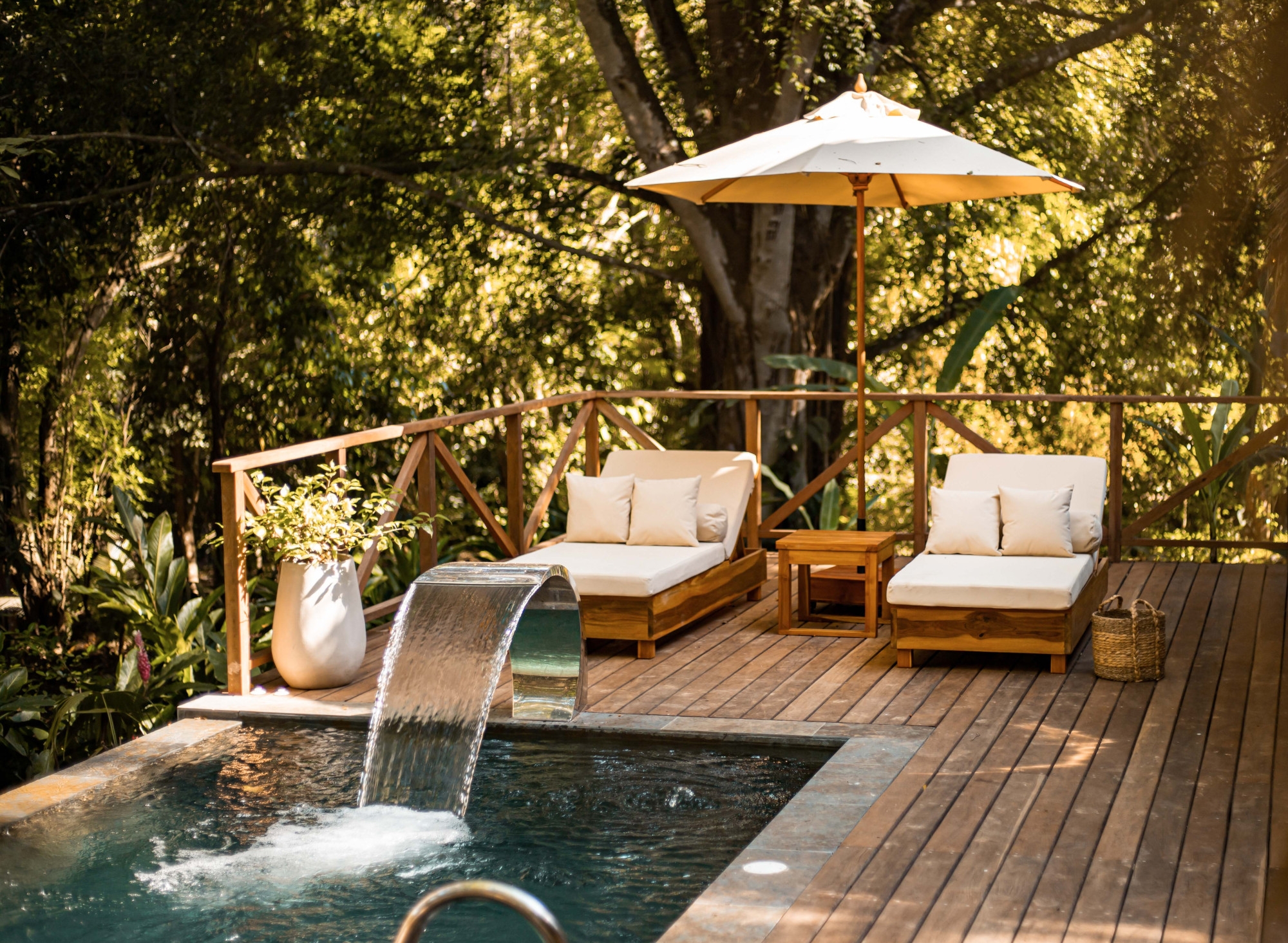 In the foreground, a small pool with a large stream of water pouring into it. Around it is a wooden deck with two white lounge chairs and a white umbrella, and in the background is dense green jungle.