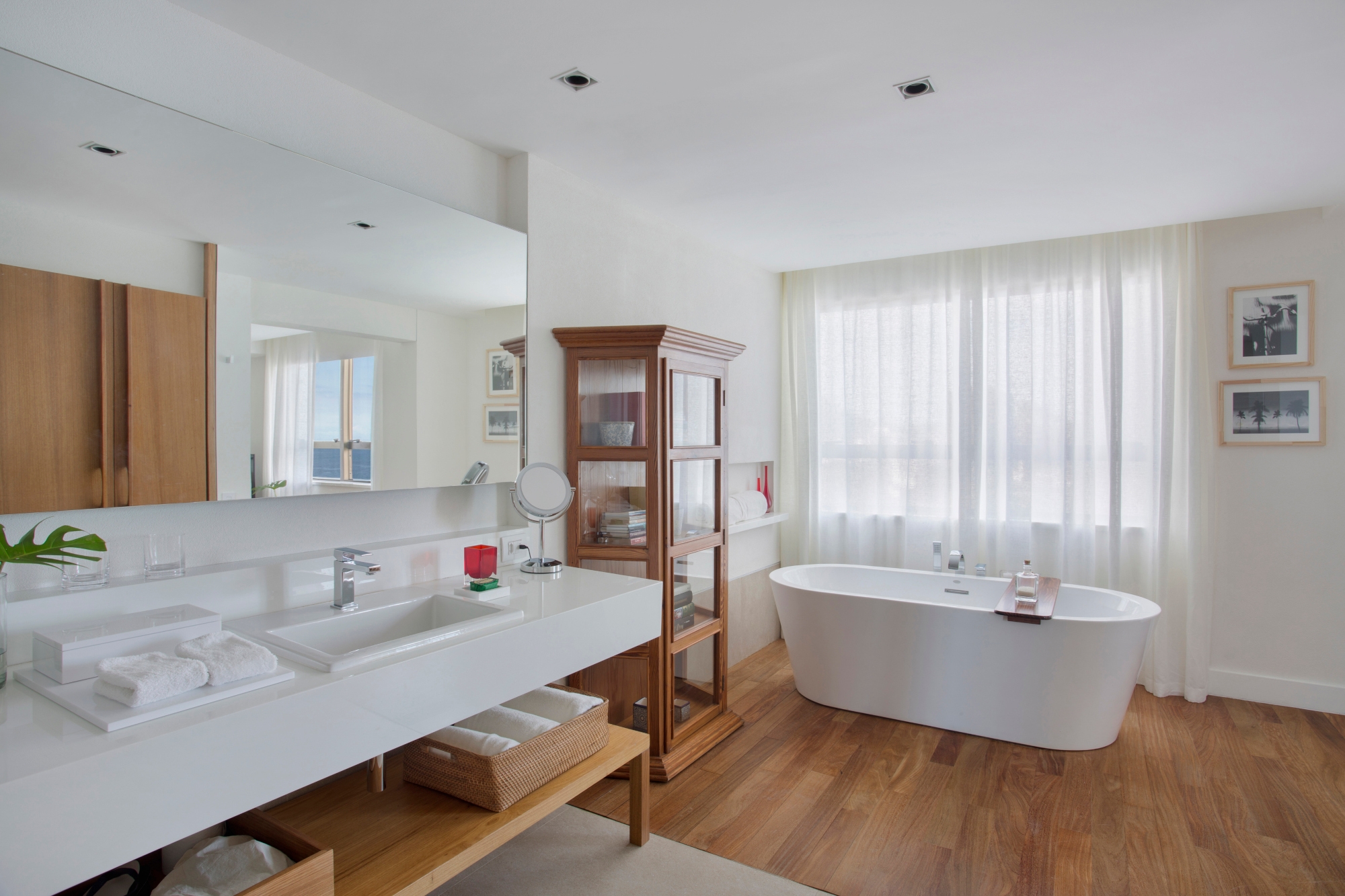 A bathroom vanity with sink and white counter underneath and next to which are light wood shelves and drawers full of towels and toilet paper. Over the sink is a mirror depicting a bedroom and to the right is a standalone white bathtub with a window and sheer curtains behind it. It stands on hardwood floors with white walls and ceiling around it.