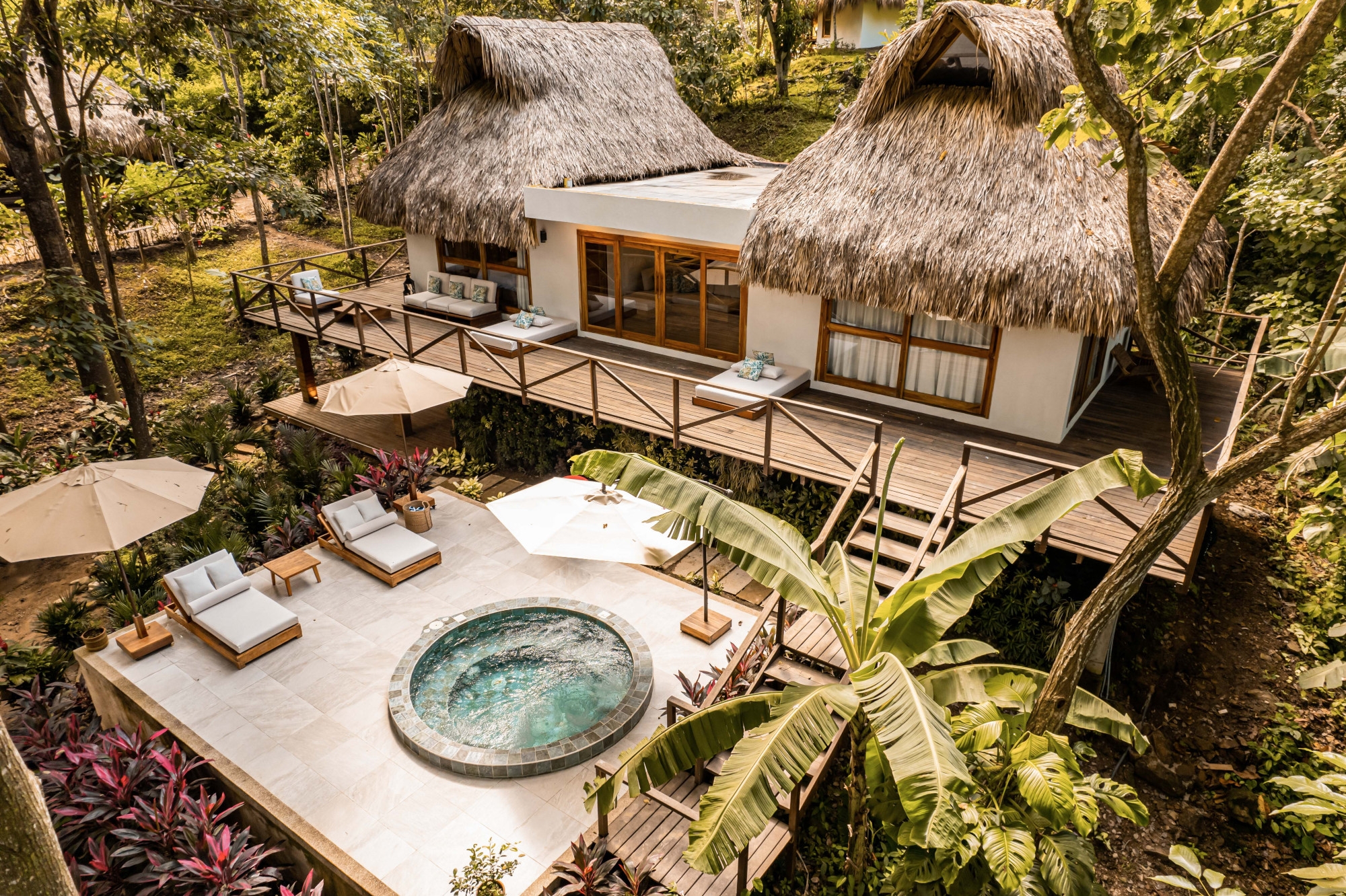 A view from above of a two-storey room with whitewashed walls, wooden railings and stairs and a thatched roof. Around it in the foreground is a deck with white lounge chairs and a small round hot tub with blue water. In the background is dense green foliage and large palm trees.