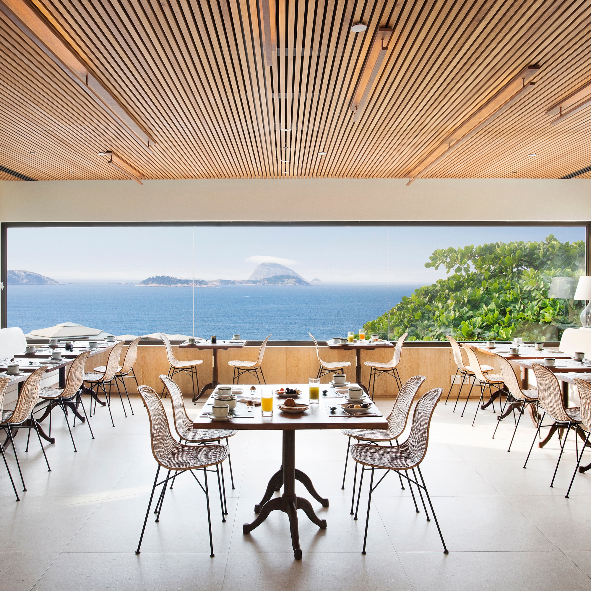 A dining room with rattan chairs and tables under a wooden ceiling and with a white floor. In the background is a long picture window looking out over some greenery and in the distance, the blue sea, some islands and the blue sky.