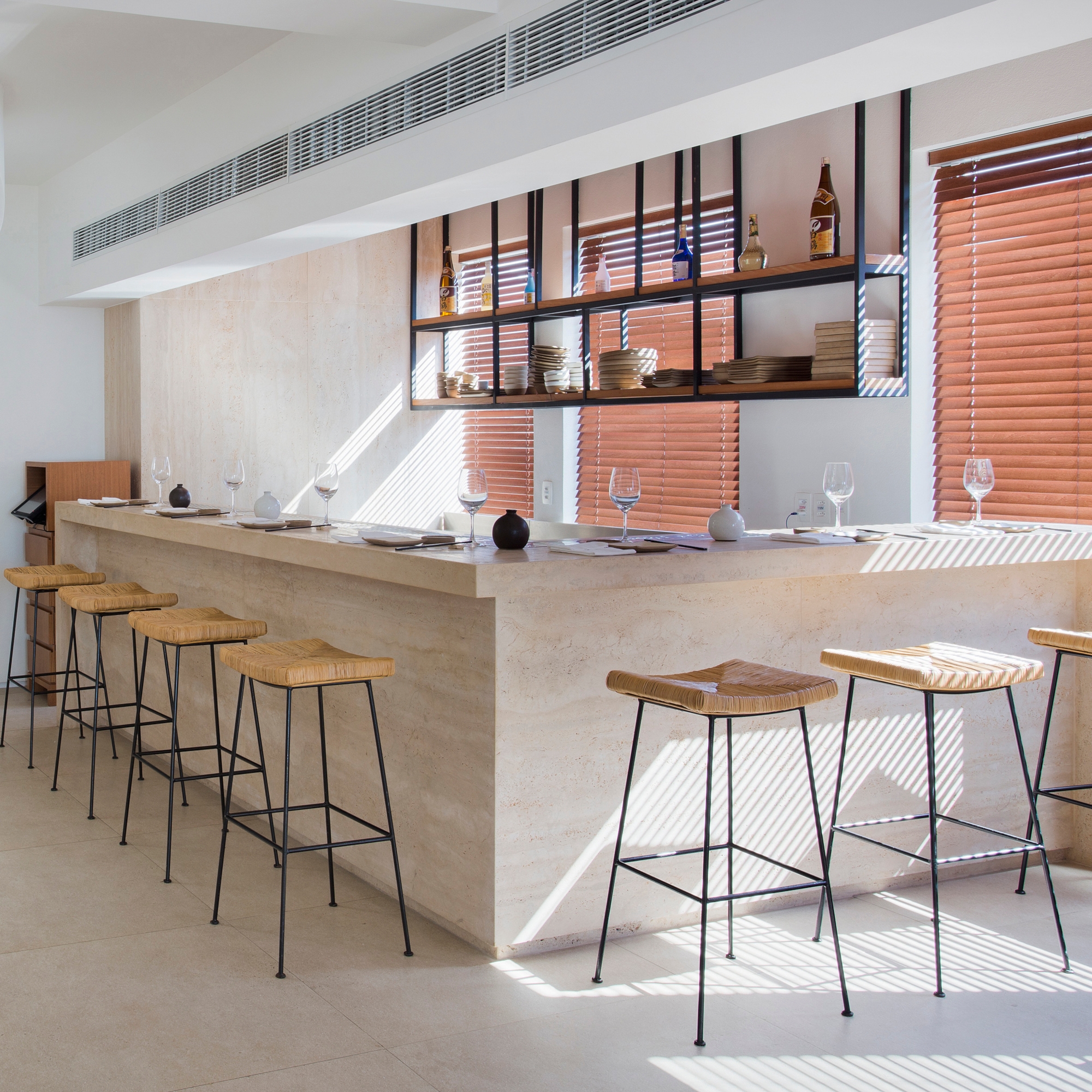 A white marble bar set with wine glasses, plates and napkins, with light wood and metal stools around two sides. Over it is a open shelf with various bottles and dinnerware. In the background is a set of three windows with dark wooden blinds and sun is shining in onto some of the stools.