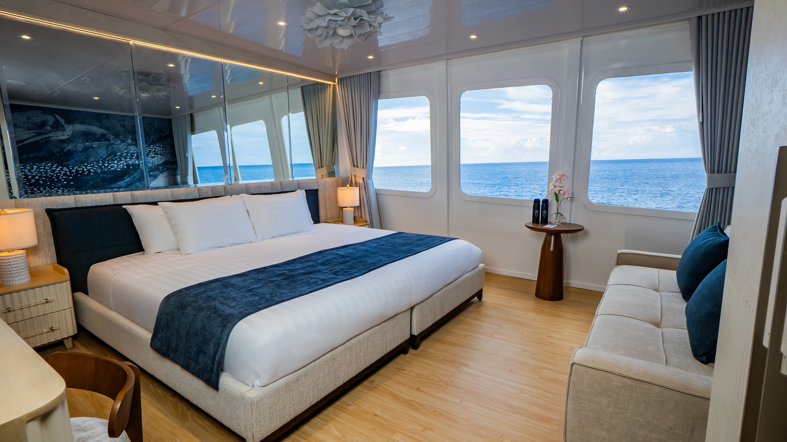 A spacious yacht bedroom with a king-size bed, mirrored headboard wall, sofa, and three large windows overlooking the sea.