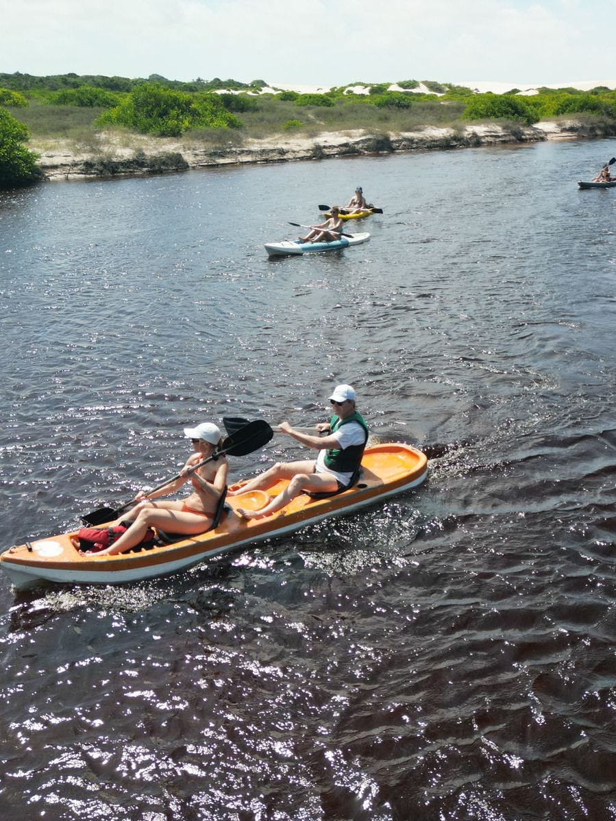 Two people in white hats row a small boat along a dark body of water, with three more boats in the background and a shoreline with white sand and grass.