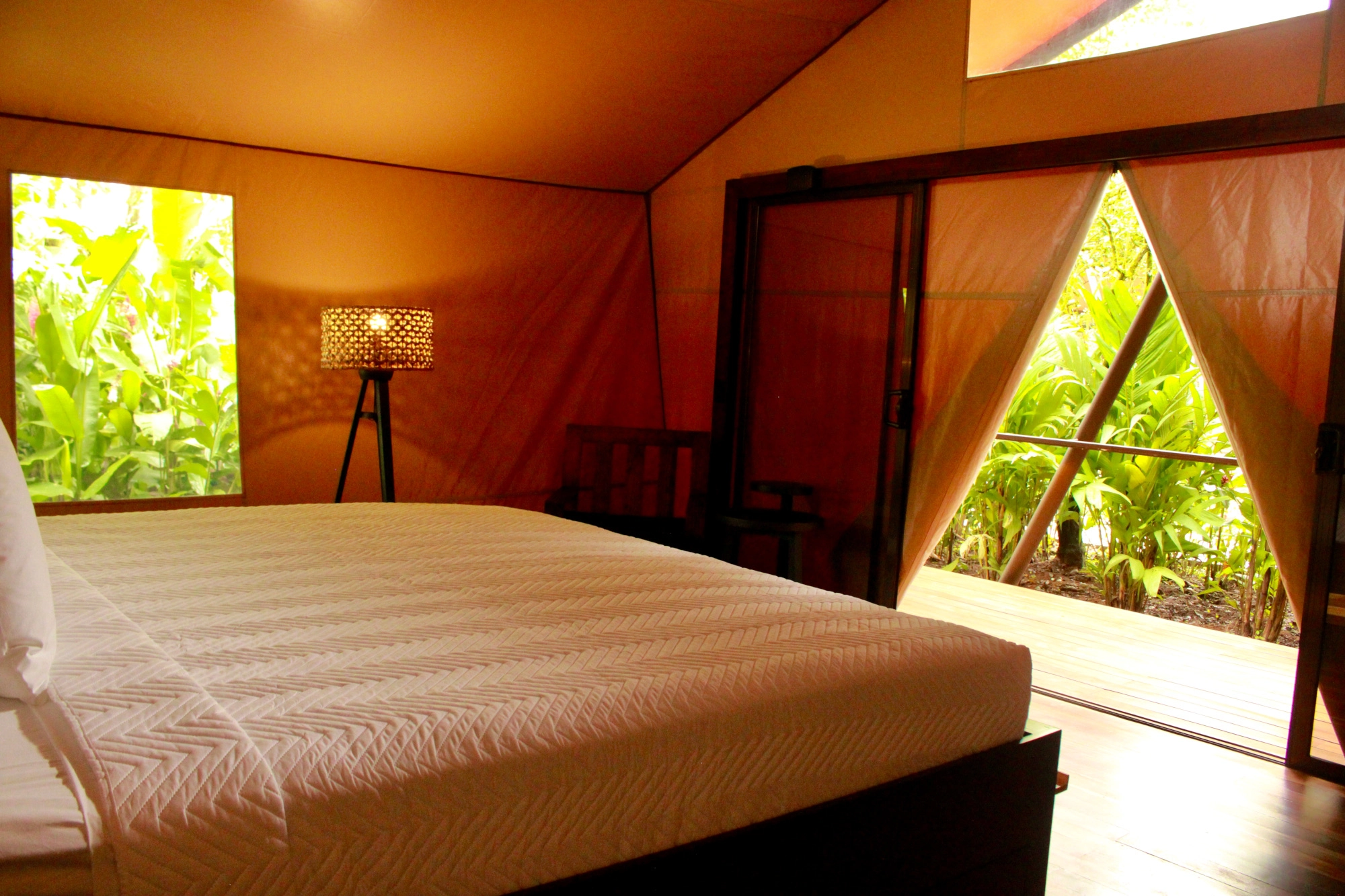 An orange-hued bedroom inside a canvas tent room with a bed with white sheets and pillows. In the background there is a lamp and a window overlooking green trees and a large door opens out onto a deck with more green trees.