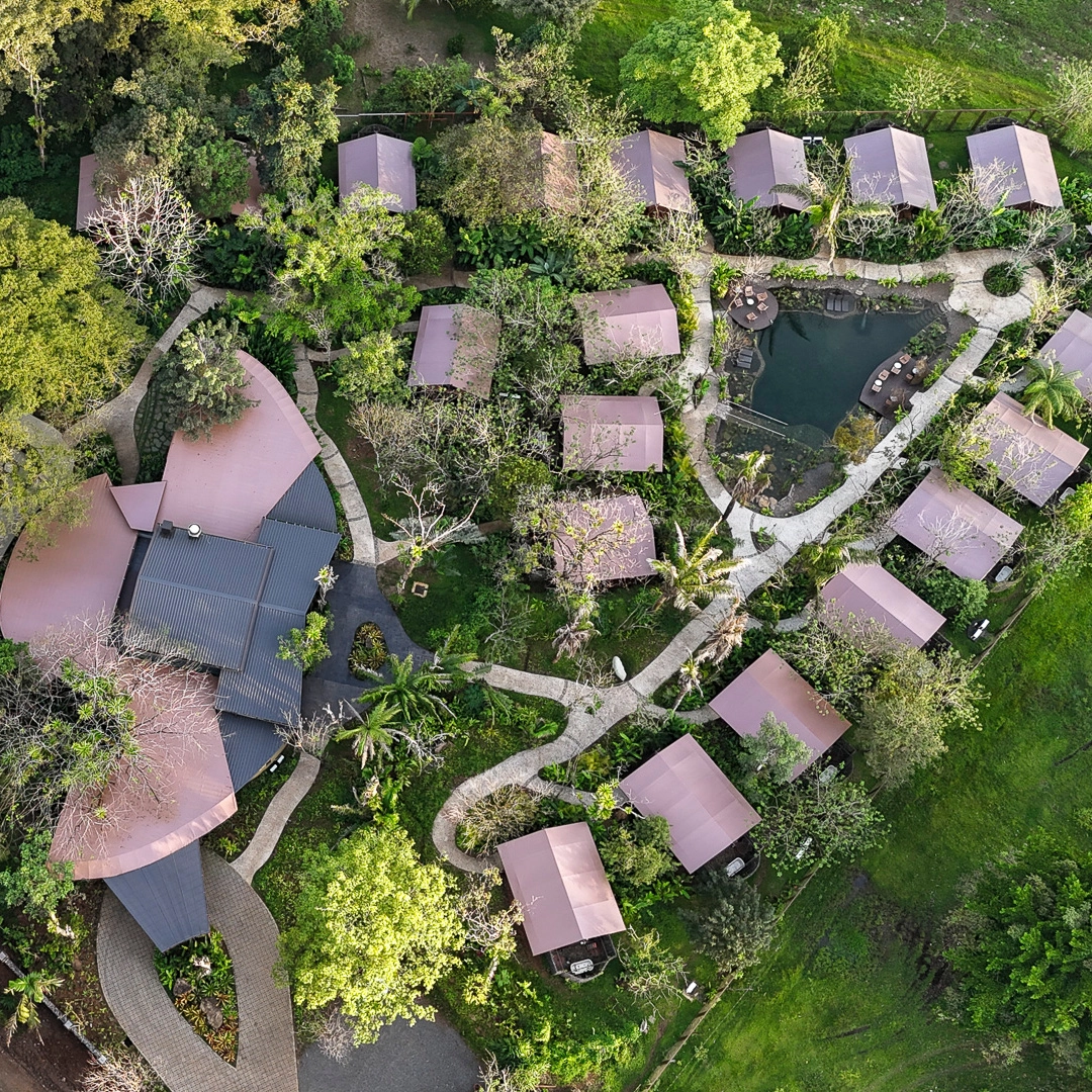 An aerial shot of a hotel with a main lodge building on the left and 18 smaller buildings can be seen to the right with some small walkways connecting them and lots of green trees and grass in between them.
