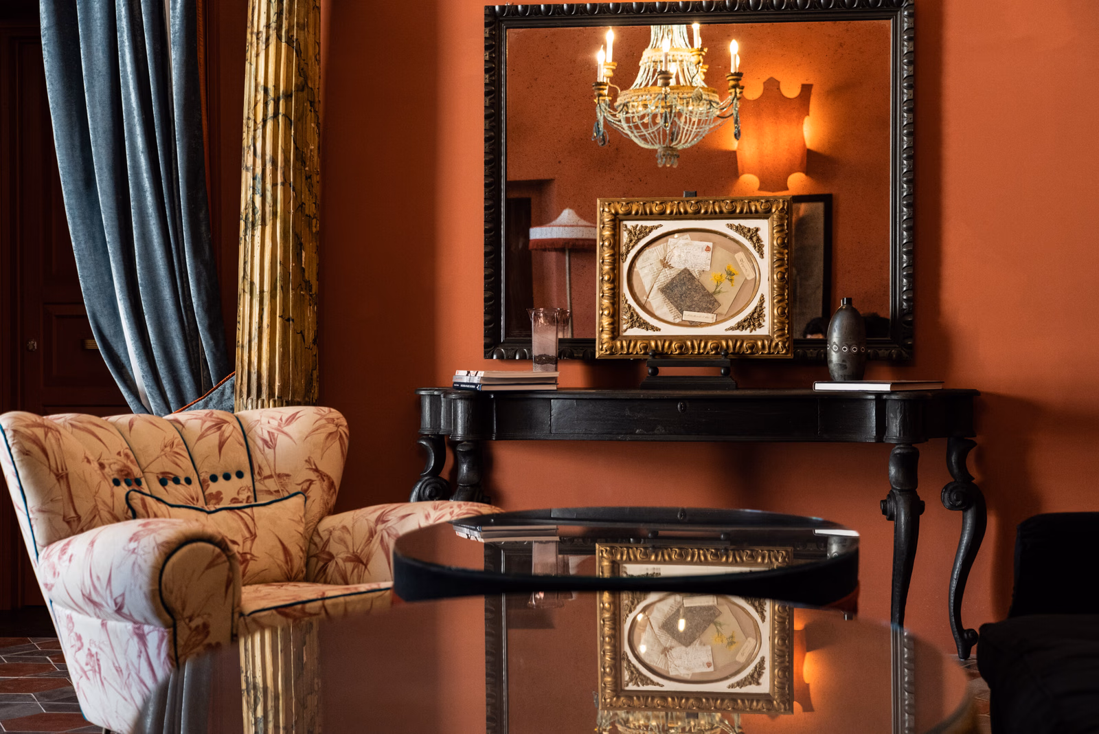 Interior shot of a lounge chair and table in a room with red walls and a big mirror reflecting a chandelier.
