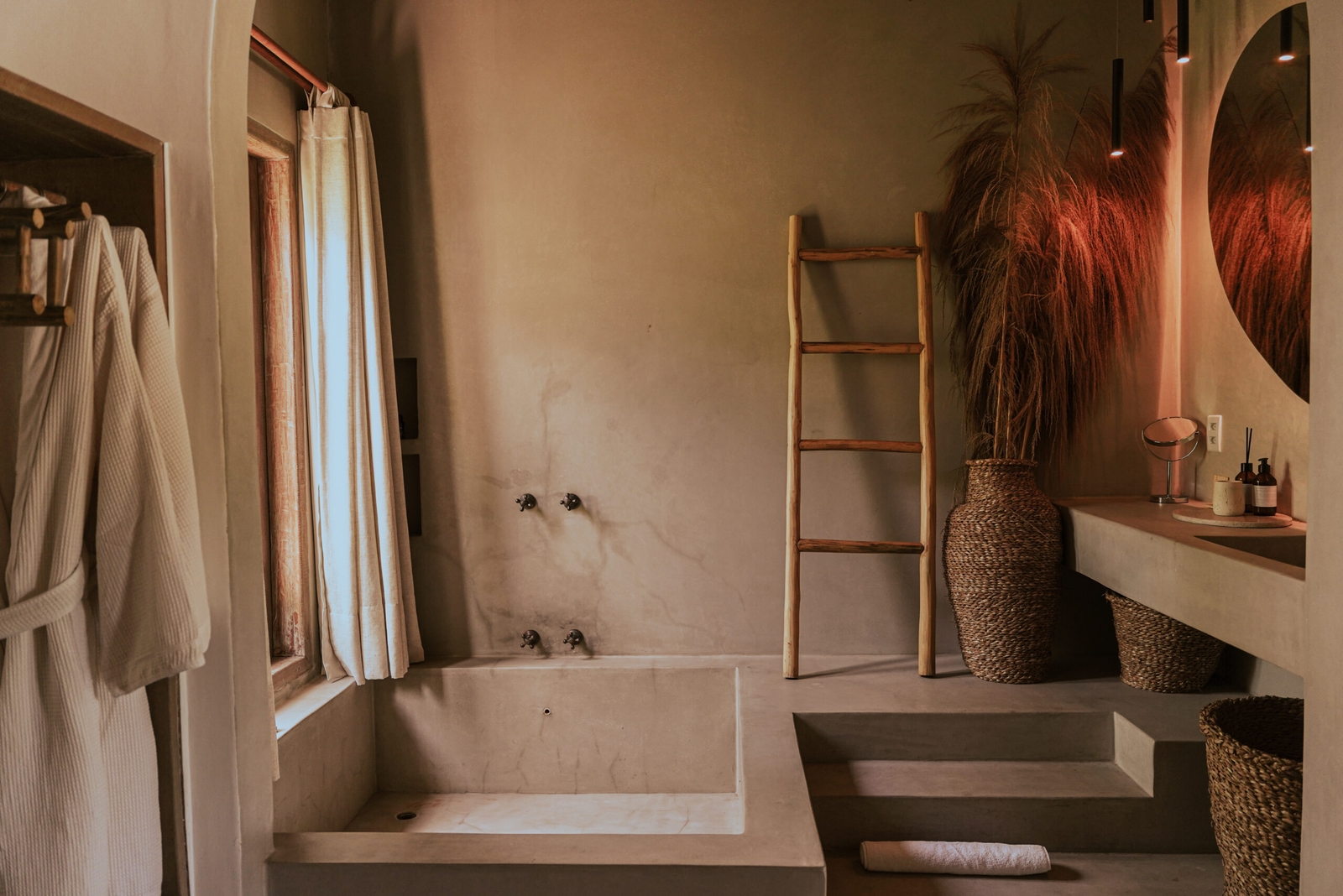 A low-lit bathroom with beige walls and a big window on the left letting in light. There is a small stone soaking tub on the left, with some steps up to it, and on the right a wooden decorative ladder and ceramic vase with a fern.